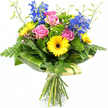 The photograph shows a vibrant mixed bouquet, hand-tied and standing upright against a pure white background, its colours appearing particularly vivid and crisp. At the centre, fully open pink roses reveal soft, layered petals that curl outwards in gentle spirals, giving the arrangement a romantic and welcoming heart. Tucked around them are bright yellow gerbera daisies, their wide petals radiating out from deep brown centres that lend a playful, almost sunburst effect, reminiscent of cheerful market days in Epping town centre. Reaching above the main cluster are tall stems of deep blue delphinium, each adorned with multiple small blossoms that create elegant vertical lines and a refreshing cool contrast to the warm pinks and yellows below. The bouquet is richly padded with varied greenery: feathery fern fronds extend from the lower edges in light, airy sprays, while broader, glossy dark green leaves sit closer to the blooms, providing a smooth, polished backdrop. Interspersed throughout are dense patches of vivid, moss-like filler foliage in lively greens, adding texture, volume, and a sense of lush abundance, as if freshly gathered from a well-tended garden just beyond Epping Forest. A few slender, pale green stems tipped with tiny buds rise gently over the flowers, hinting at new blossoms to come. At the base of the display, large arched leaves with subtle lighter green striping are wrapped around the stems in a tidy collar that frames the bouquet. Beneath this natural collar, the neatly aligned stems are bound together in a soft, pale wrap with a simple tie, clearly showing its hand-tied craftsmanship. The arrangement feels joyful and uplifting, ideal for gifting or local flower delivery around Epping to mark birthdays, congratulations, or everyday moments of appreciation.